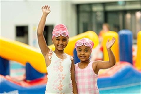 Two children in swimwear waving in front of a swimming pool with large infatables in it.